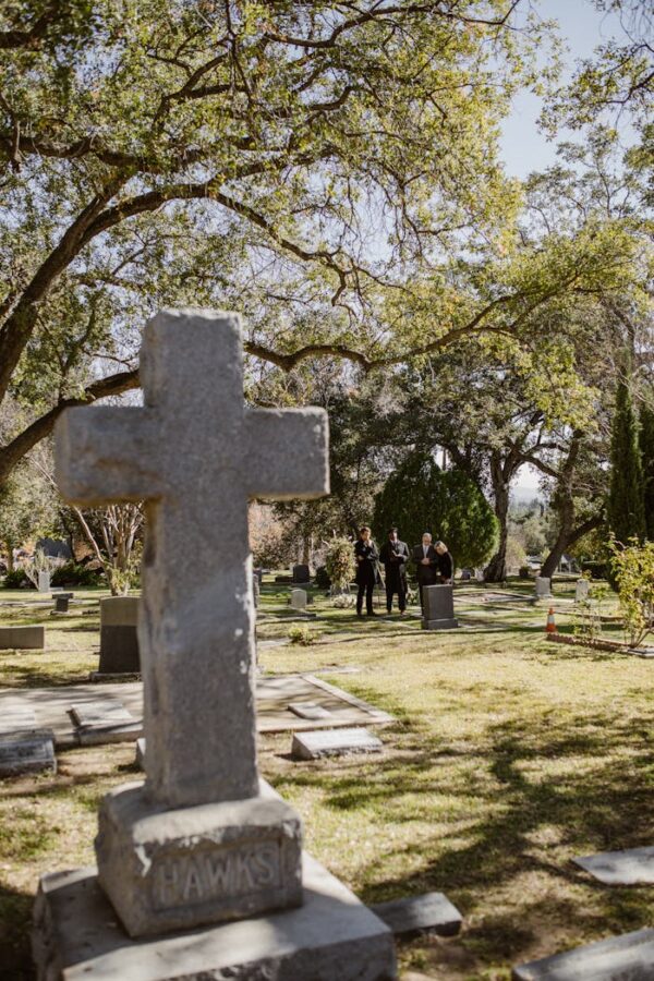 pexels-photo-6841451 A serene cemetery with a prominent stone cross surrounded by lush greenery and gravestones.